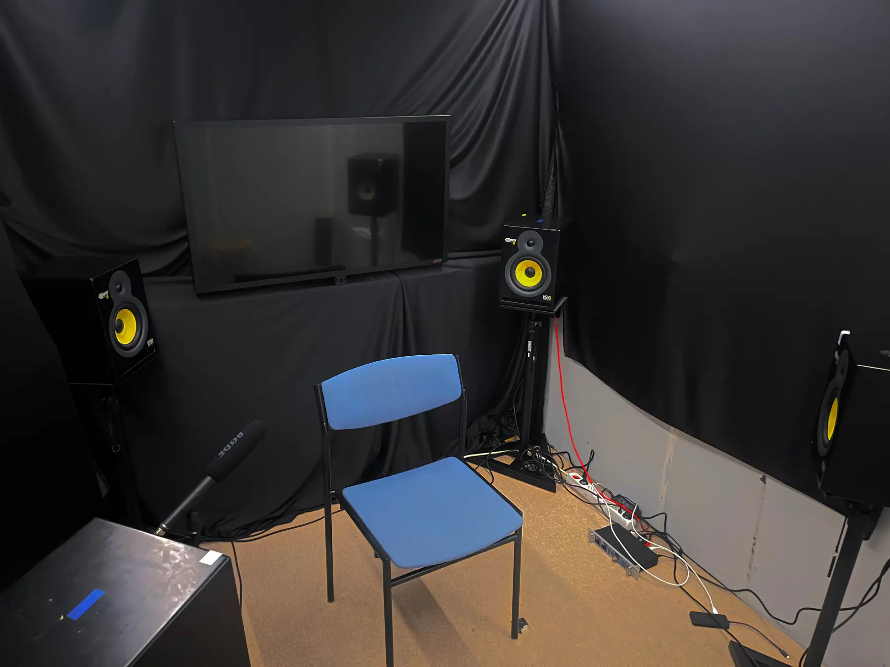 A blue chair in a listening booth surrounded by studio monitors and black acoustic curtains.