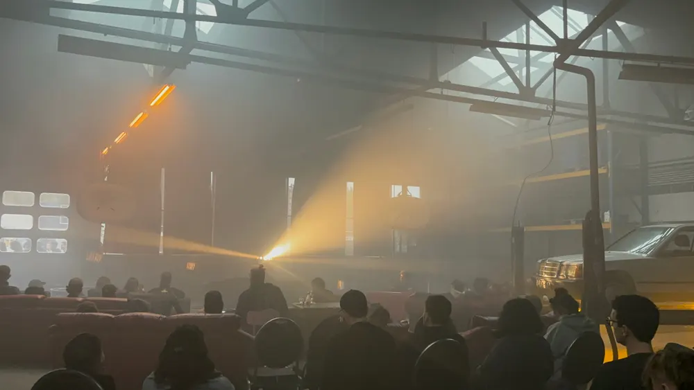 An audience in a hazy warehouse lit by stage lights.