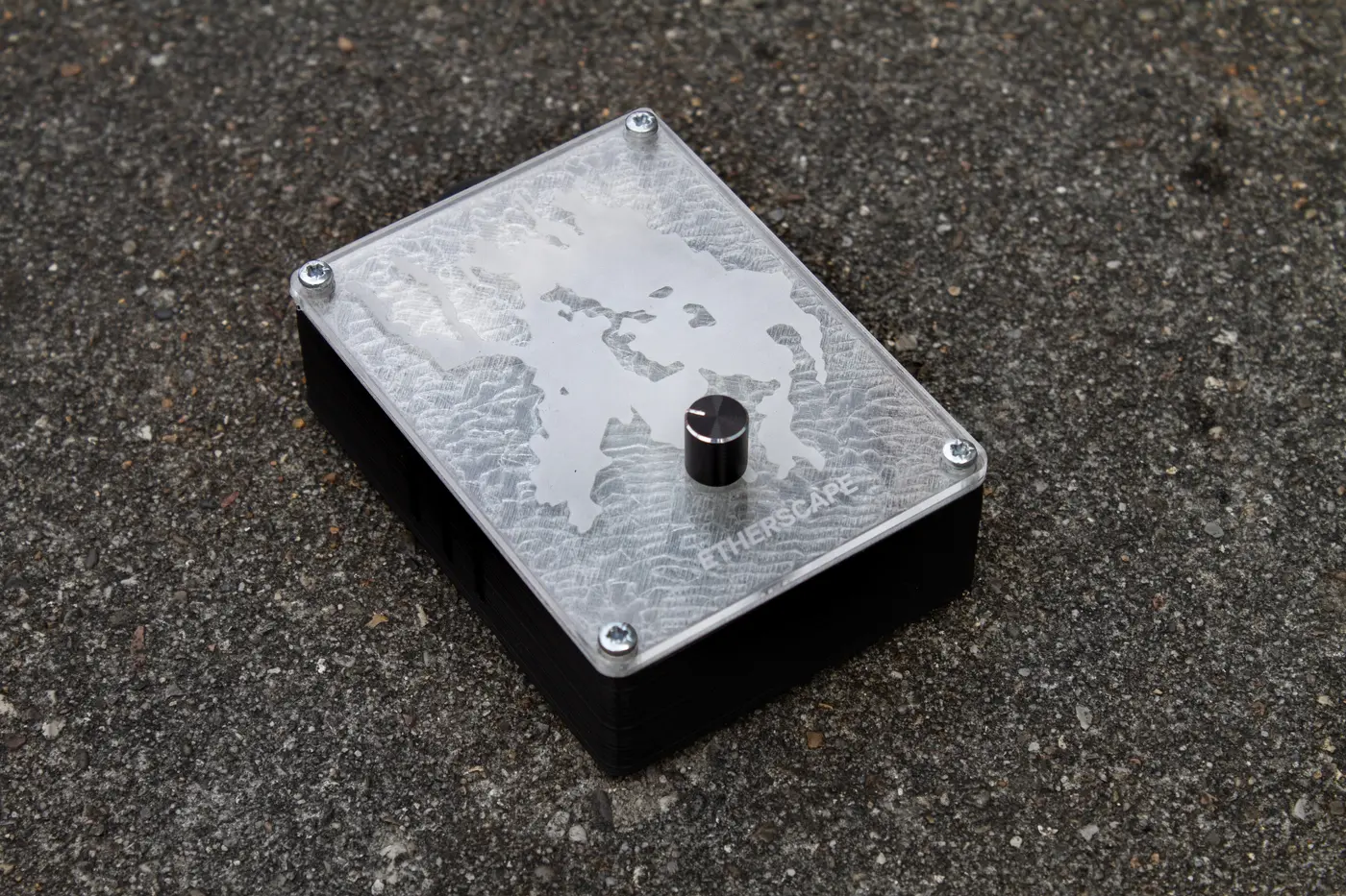A small black box with an etched acrylic top and a single knob, resting on asphalt.
